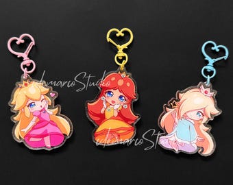 Puede incluir: Tres llaveros acrílicos con princesas de dibujos animados. La Princesa Peach con un vestido rosa, la Princesa Daisy con un vestido amarillo y la Princesa Rosalina con un vestido azul. Cada llavero tiene un cierre en forma de corazón.