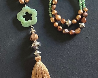 Op de afbeelding: Een lange ketting met bruine en groene kralen, een jadegroene bloempendant en een bruine kwast.
