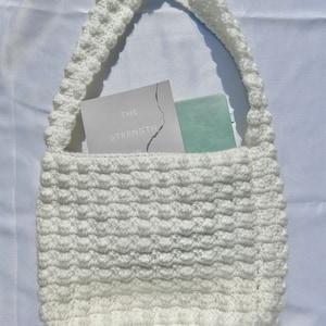 May include: White crocheted tote bag with a thick, textured strap. The bag is filled with a book titled "The Strength In" and a light blue rectangle.
