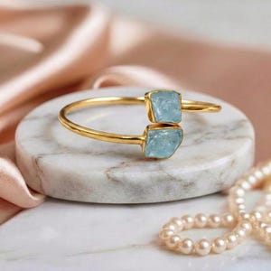May include: A gold-colored bracelet with two light blue, square-shaped gemstones. The bracelet is resting on a white and gray marble surface, with a pink satin fabric in the background. A string of pearls is in the foreground.