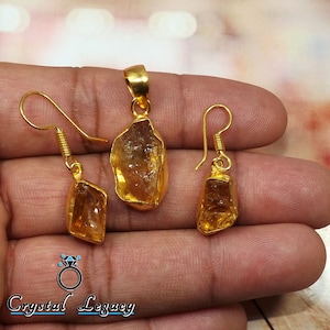 Pu&ograve; includere: Un set di gioielli color oro con un ciondolo e orecchini. Ogni pezzo presenta una pietra preziosa grezza color ambra incastonata in oro. Gli orecchini hanno un design a gancio.