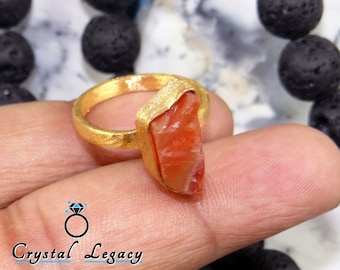 May include: A gold-toned ring featuring a rough-cut, reddish-orange gemstone. The ring has a textured band and a unique setting that cradles the stone. The image also includes the text "Crystal Legacy".