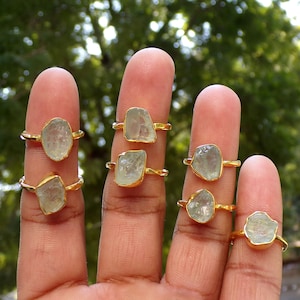 Könnte beinhalten: Acht goldene Ringe mit klaren, facettierten, rohen Aquamarin-Steinen. Die Ringe werden an den Fingern einer Hand getragen.