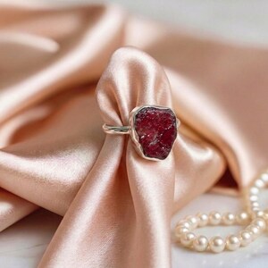 May include: A silver ring featuring a large, irregularly shaped, deep red gemstone. The ring is set against a backdrop of soft, peach-colored fabric and a strand of pearls, highlighting the jewelry's elegant design.