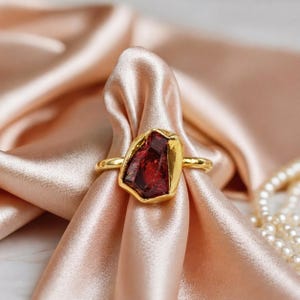 Op de afbeelding: Een gouden ring met een grote, onregelmatig gevormde, dieprode edelsteen. De ring is geplaatst tegen een achtergrond van zachte, perzikroze stof, met een parelsnoer zichtbaar. De ring heeft een eenvoudige, gepolijste gouden band.