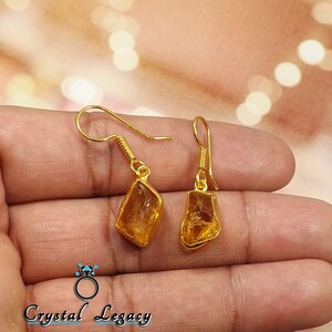 Pu&ograve; includere: Un paio di orecchini color oro con pendenti di pietre preziose grezze color ambra. Gli orecchini hanno un design a gancio e sono mostrati su uno sfondo sfocato. Il testo "Crystal Legacy" &egrave; in basso.