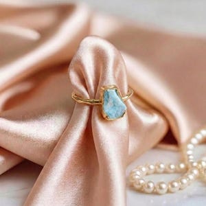 Könnte beinhalten: Ein goldfarbener Ring mit einem hellblauen, unregelmäßig geformten Stein. Der Ring ist vor einem Hintergrund aus weichem, rosa Stoff und einer Perlenkette platziert, was eine elegante Ästhetik erzeugt. Das Design des Rings ist schlicht, aber auffällig.