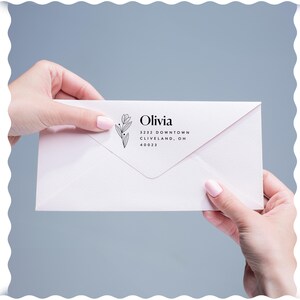 Może przedstawiać: Biała koperta z imieniem "Olivia" i danymi adresowymi wydrukowanymi na czarno. Po lewej stronie imienia znajduje się kwiatowy wzór. Koperta jest trzymana na jasnoniebieskim tle.