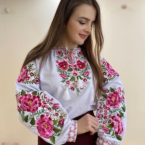 Könnte beinhalten: Weiße bestickte Bluse mit rosa und grünen Blumenmustern. Die Bluse hat einen runden Ausschnitt und lange Ärmel.