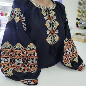 May include: A black embroidered blouse with long, puffed sleeves. The blouse features intricate floral and geometric patterns in orange, green, and white thread. The neckline and cuffs are also detailed with embroidered designs. A black tassel hangs from the neckline.