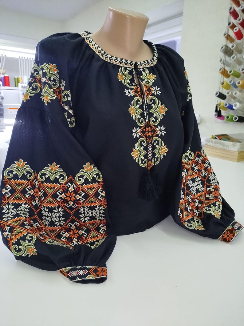 May include: A black embroidered blouse with long, puffed sleeves. The blouse features intricate floral and geometric patterns in orange, green, and white thread. The neckline and cuffs are also detailed with embroidered designs. A black tassel hangs from the neckline.