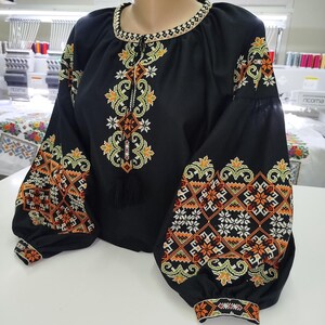 May include: Black blouse with intricate embroidery in orange, yellow, and white. The long-sleeved top features a round neckline and a tassel detail. The sleeves and front are adorned with detailed floral and geometric patterns.