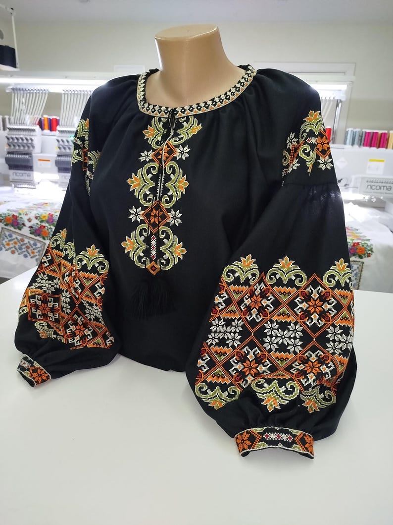 May include: Black blouse with intricate embroidery in orange, yellow, and white. The long-sleeved top features a round neckline and a tassel detail. The sleeves and front are adorned with detailed floral and geometric patterns.