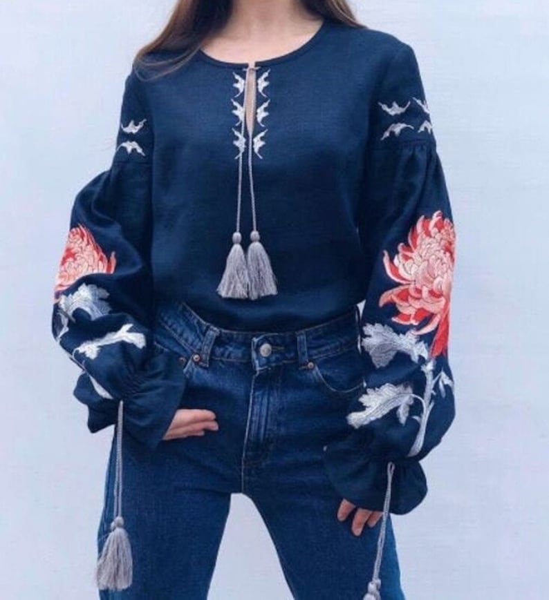 May include: A navy blue linen blouse with embroidered white and red flowers on the sleeves. The blouse has a round neckline and a tassel tie at the front.