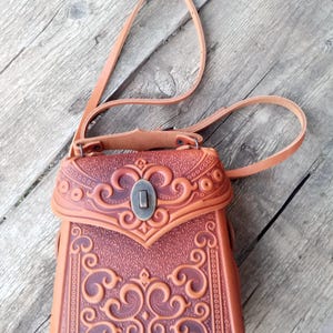 Könnte beinhalten: Eine braune Leder-Umhängetasche mit aufwendigen, geprägten Blumenmustern. Die Tasche hat einen Klappverschluss mit einer Metallschnalle und einen langen, verstellbaren Riemen.