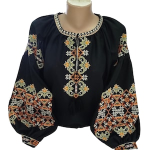 May include: Black blouse with intricate embroidery in orange, yellow, and white. The long sleeves and neckline are adorned with detailed patterns. The blouse has a round neckline and a tassel detail.