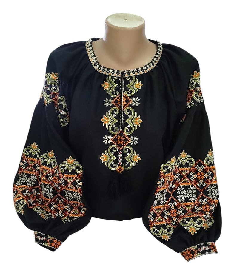 May include: Black blouse with intricate embroidery in orange, yellow, and white. The long sleeves and neckline are adorned with detailed patterns. The blouse has a round neckline and a tassel detail.