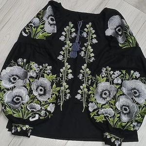 May include: Black embroidered blouse with a floral design featuring white and green flowers. The blouse has a round neckline and long sleeves.