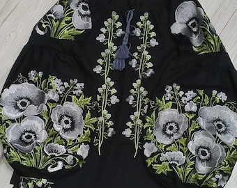 May include: Black embroidered blouse with a floral design featuring white and green flowers. The blouse has a round neckline and long sleeves.
