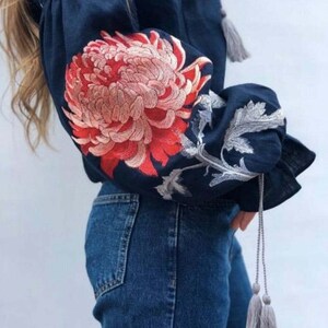May include: A navy blue linen blouse with embroidered red and white flowers on the sleeves. The blouse has a gathered neckline and long sleeves with grey tassels.