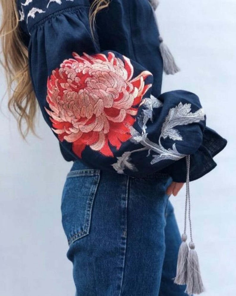 May include: A navy blue linen blouse with embroidered red and white flowers on the sleeves. The blouse has a gathered neckline and long sleeves with grey tassels.