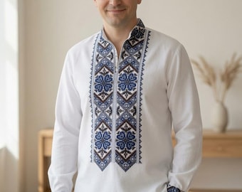 May include: A white long-sleeved shirt featuring a stand-up collar and a central vertical panel decorated with intricate blue, brown, and black geometric embroidery. The same detailed embroidery pattern is repeated on the cuffs of the sleeves.