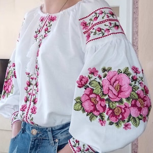 May include: White embroidered blouse with long sleeves and a floral pattern. The sleeves have a large pink rose design, while the front features a smaller floral pattern.