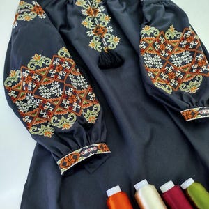 May include: A dark navy blue embroidered dress with long sleeves. The sleeves and neckline feature intricate floral and geometric patterns in orange, white, and green. A black tassel hangs from the neckline. Several spools of thread are visible.