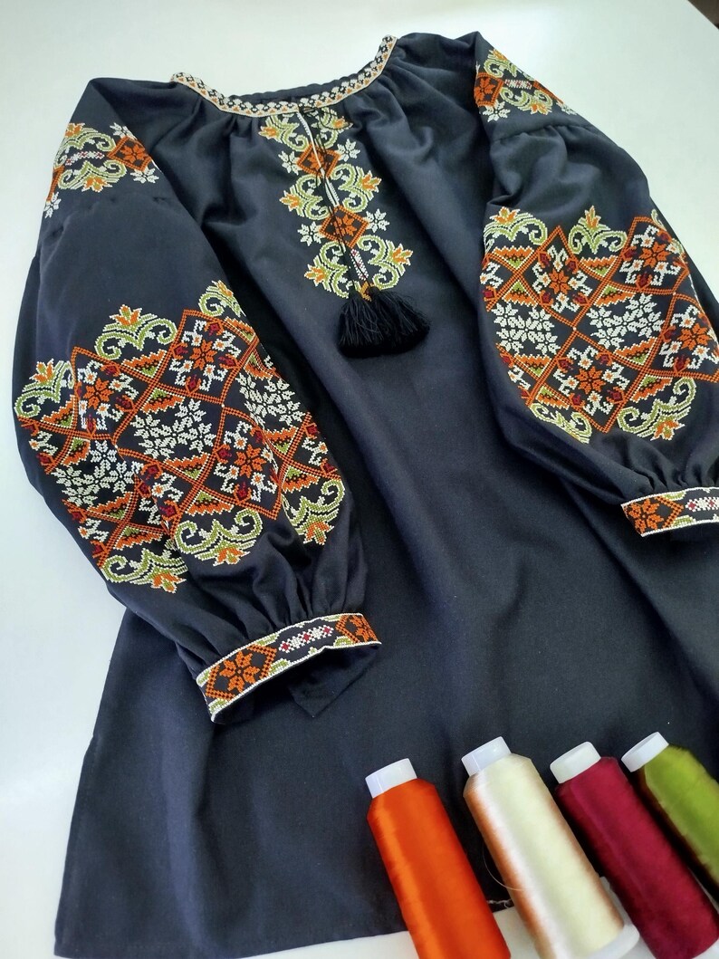 May include: A dark navy blue embroidered dress with long sleeves. The sleeves and neckline feature intricate floral and geometric patterns in orange, white, and green. A black tassel hangs from the neckline. Several spools of thread are visible.