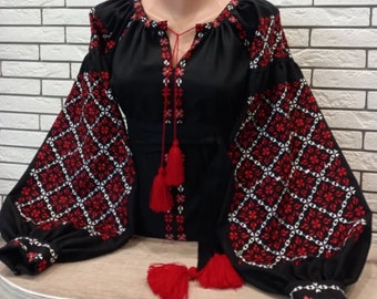 May include: Black embroidered blouse with red and white geometric patterns on the sleeves and neckline. The blouse has a long sleeve and a loose fit.