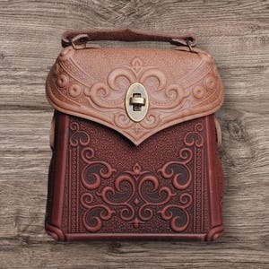 Op de afbeelding: Een bruine leren handtas met ingewikkelde gegraveerde bloemmotieven. De tas heeft een klep sluiting met een metalen sluiting en een korte band.