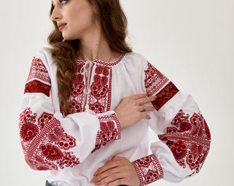 May include: White blouse with long sleeves and intricate red embroidery. The blouse features detailed floral patterns on the sleeves, chest, and cuffs. The model is wearing blue jeans and a black belt.