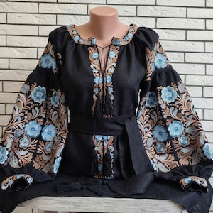 May include: Black embroidered blouse with blue floral patterns. The blouse has a tie-up neckline, a matching belt, and tassels. The long, puffy sleeves have embroidered cuffs. The embroidery includes shades of blue and brown.