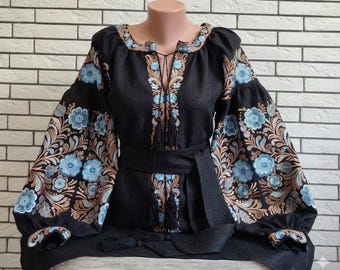 Könnte beinhalten: Schwarze bestickte Bluse mit blauen Blumenmustern auf den Ärmeln und dem Oberteil. Die Bluse hat einen Schnürhalsausschnitt, einen passenden Gürtel und dekorative Quasten. Die Ärmel sind lang und puffig, mit bestickten Manschetten.