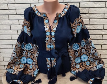 Op de afbeelding: Een marineblauwe linnen blouse met geborduurde bloemenpatronen in goud en lichtblauw. De blouse heeft een V-hals, lange mouwen en een strik in de taille.