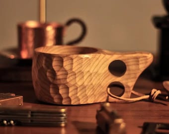 May include: A wooden cup with a unique, textured design. The cup has two holes on the side, one of which has a leather strap attached.