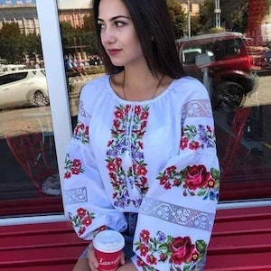Może przedstawiać: Biała haftowana bluzka z czerwonymi, różowymi i fioletowymi wzorami kwiatowymi. Długie rękawy zdobią misterne koronkowe detale i duże czerwone róże. Bluzka jest połączona z dżinsowymi szortami. W dłoniach trzymany jest czerwony kubek z kawą.