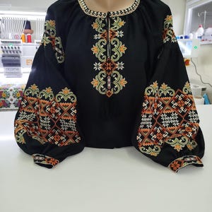 May include: Black blouse with intricate floral embroidery in orange, green, and white. The long-sleeved top features a round neckline and a central embroidered design. The sleeves have a puffed design and are also embroidered.
