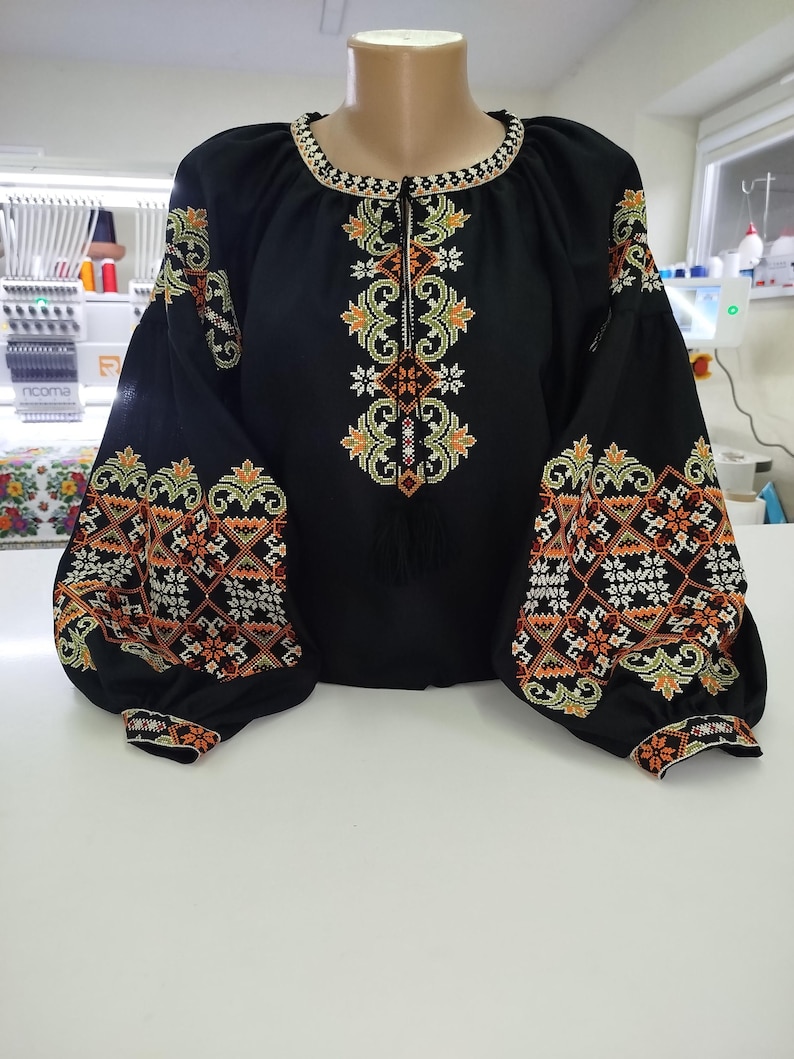 May include: Black blouse with intricate floral embroidery in orange, green, and white. The long-sleeved top features a round neckline and a central embroidered design. The sleeves have a puffed design and are also embroidered.