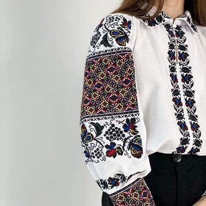 Puede incluir: Blusa blanca con intrincados bordados florales en rojo, azul, amarillo y negro. Las mangas largas y el cuello presentan patrones detallados. La blusa se combina con pantalones negros.