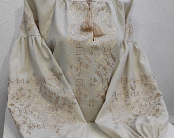 May include: A beige linen blouse with embroidered geometric patterns in a light gold color. The blouse has long sleeves with gathered cuffs and a round neckline.