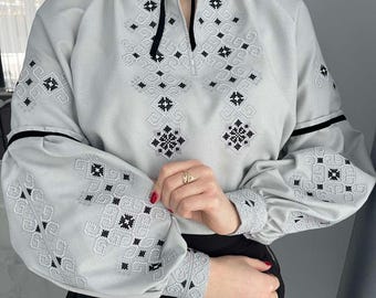 May include: A light gray blouse with long sleeves and intricate black embroidery. The blouse features a black ribbon tie at the neckline and black trim on the sleeves. The sleeves are gathered at the wrists.