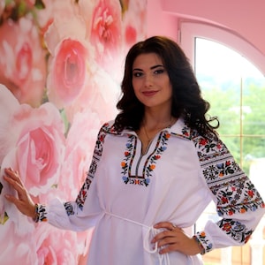 Puede incluir: Una mujer lleva una blusa blanca bordada con una faja blanca y pantalones negros. La blusa tiene bordados florales coloridos en las mangas y el escote. La mujer está de pie frente a una pared con un patrón floral.