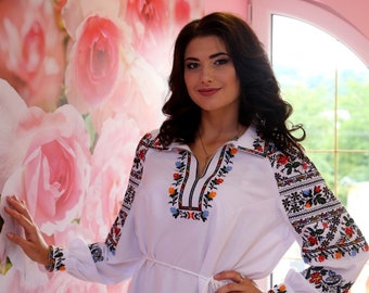 May include: A woman wearing a white embroidered blouse with a white sash and black pants. The blouse has colorful floral embroidery on the sleeves and neckline. The woman is standing in front of a wall with a floral pattern.