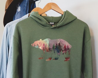 May include: A green hooded sweatshirt with a colorful bear silhouette design. The bear is filled with a watercolor-like pattern of trees and a sunset. The text "PLUS SIZES" is on a brown circle in the lower right corner of the image.