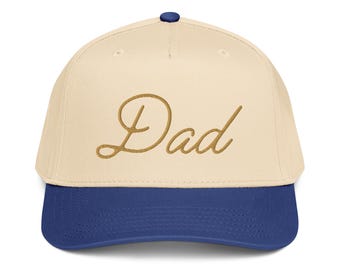 Op de afbeelding: Een baseballpet met een crèmekleurige kroon en een marineblauwe rand. Het woord "Dad" is in een gouden script over de voorkant van de pet geborduurd. De pet heeft een marineblauwe knoop bovenop.