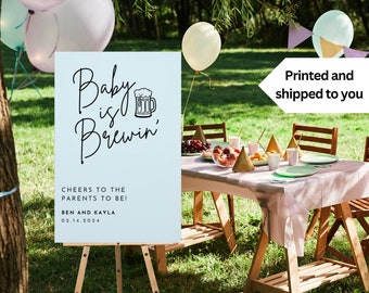 May include: A light blue baby shower sign with the text "Baby is Brewin'" and a black and white illustration of a beer mug. The sign also includes the text "Cheers to the parents to be! Ben and Kayla 02.14.2024".