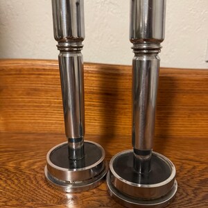 May include: Two silver-colored candle holders with a classic design. Each holder has a cylindrical body, a flared top, and a circular base with a black center. The candle holders are displayed on a wooden surface.