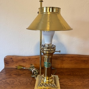 May include: A vintage brass desk lamp with a conical shade and ornate base. The lamp features a decorative finial and a label on the central column. The lamp is plugged into an electrical outlet.