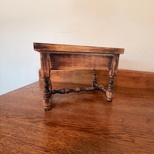 May include: A small wooden stand with a rectangular top and four turned legs connected by a lower cross-stretcher. The wood features a dark, distressed finish with visible grain patterns and darkened edges, creating a weathered appearance.